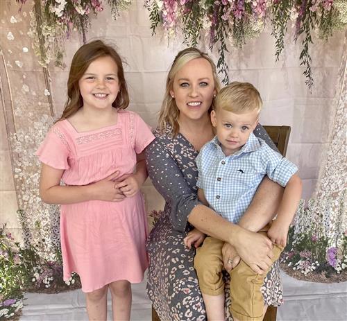 Keri Lynn McDonald smiles to camera while sitting down. She has her son in her lap and her daughter standing next to her. 